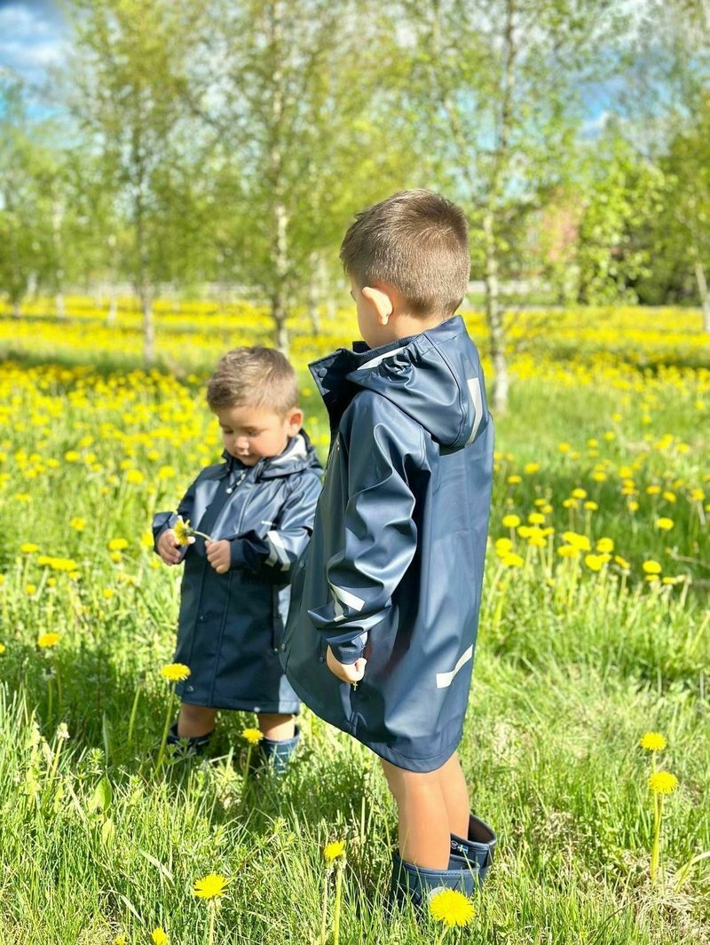 To børn har mørkeblå vandtætte regnjakker og gummistøvler på. Omgivelserne har grønt græs og gule mælkebøtter under en blå himmel.