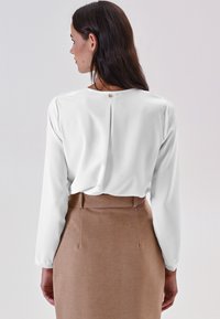 Donna con lunghi capelli scuri che indossa una camicetta bianca a maniche lunghe infilata in una gonna beige alta in vita, di spalle rispetto alla fotocamera.