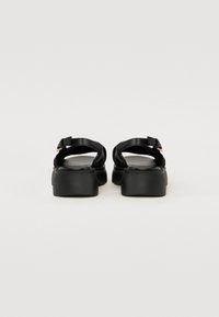 Mustat sandaalit paksulla pohjalla, joissa on punottu hihna ja säädettävä nilkkahihna. Tasainen nahkapinta.