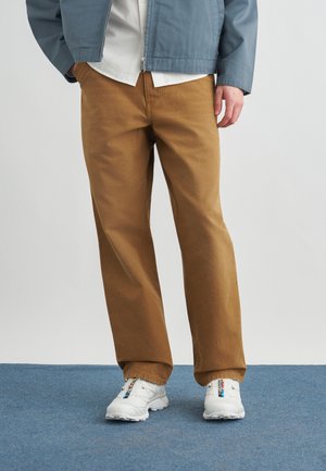 Osoba w brązowych spodniach o prostych nogawkach, białych tenisówkach z kolorowymi detalami, niebieskiej kurtce i białej koszuli stoi na niebieskim dywanie.