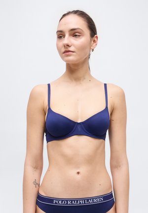 Mujer con sujetador azul marino y ropa interior a juego con el logo de Polo Ralph Lauren en la cintura, de pie contra un fondo claro y liso.
