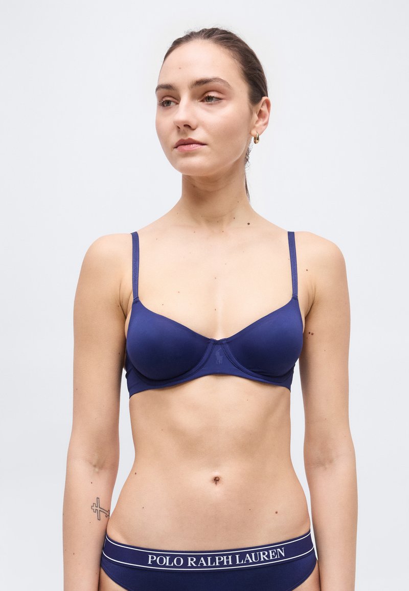 Γυναίκα που φοράει μπλε σκούρο σουτιέν και ταιριαστό εσώρουχο με το λογότυπο Polo Ralph Lauren στη ζώνη, στέκεται μπροστά σε ένα απλό φωτεινό φόντο.