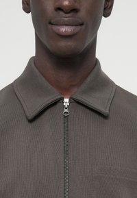 Γκρι μπουφάν με φερμουάρ, επίπεδη γιακά, υφασμάτινη υφή και τσέπη στο στήθος, με ασημένιο φερμουάρ και διακριτικές ραμμένες λεπτομέρειες.