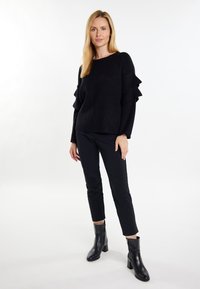 Mustardärinen neulepaita, jossa on röyhelöhihoja, yhdistettynä slim fit -mustiin housuihin ja nilkkureihin. Silkkinen pinta ja rento leikkaus.