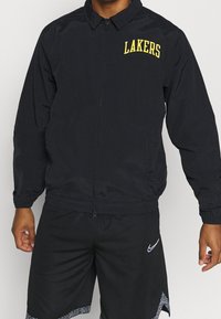 Schwarze Zip-Jacke mit Kragen, mit "LAKERS" in Gelb auf der linken Brust. Windbreaker-Material, leichtes Design.