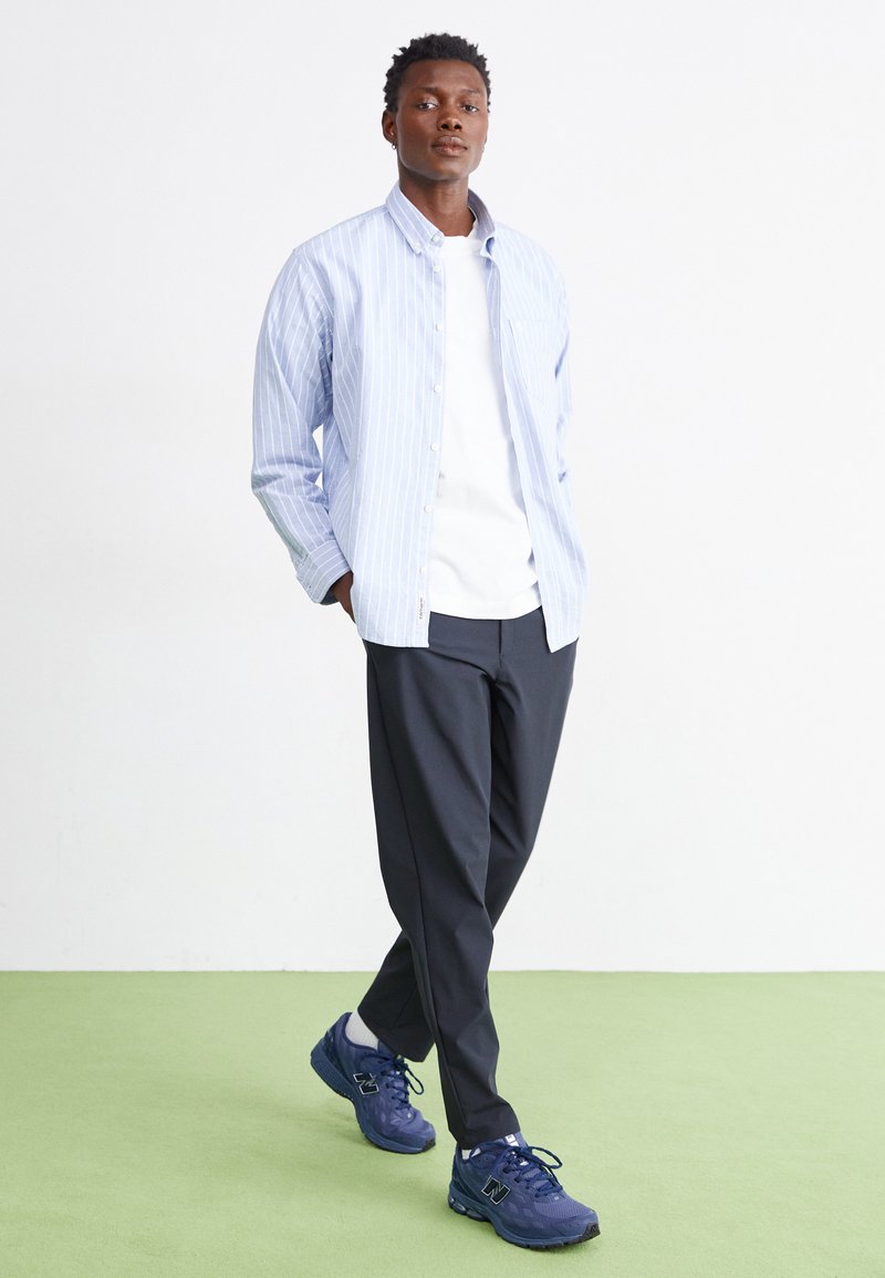 Chemise boutonnée rayée bleu clair portée sur un t-shirt blanc, associée à un pantalon gris foncé et des chaussures de course bleues navy. Se tenant sur un sol vert.