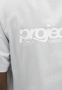 Lys grå bomulds t-shirt med et stort hvidt logo med teksten "projec" og "CREATIVE." Glat tekstur, korte ærmer.