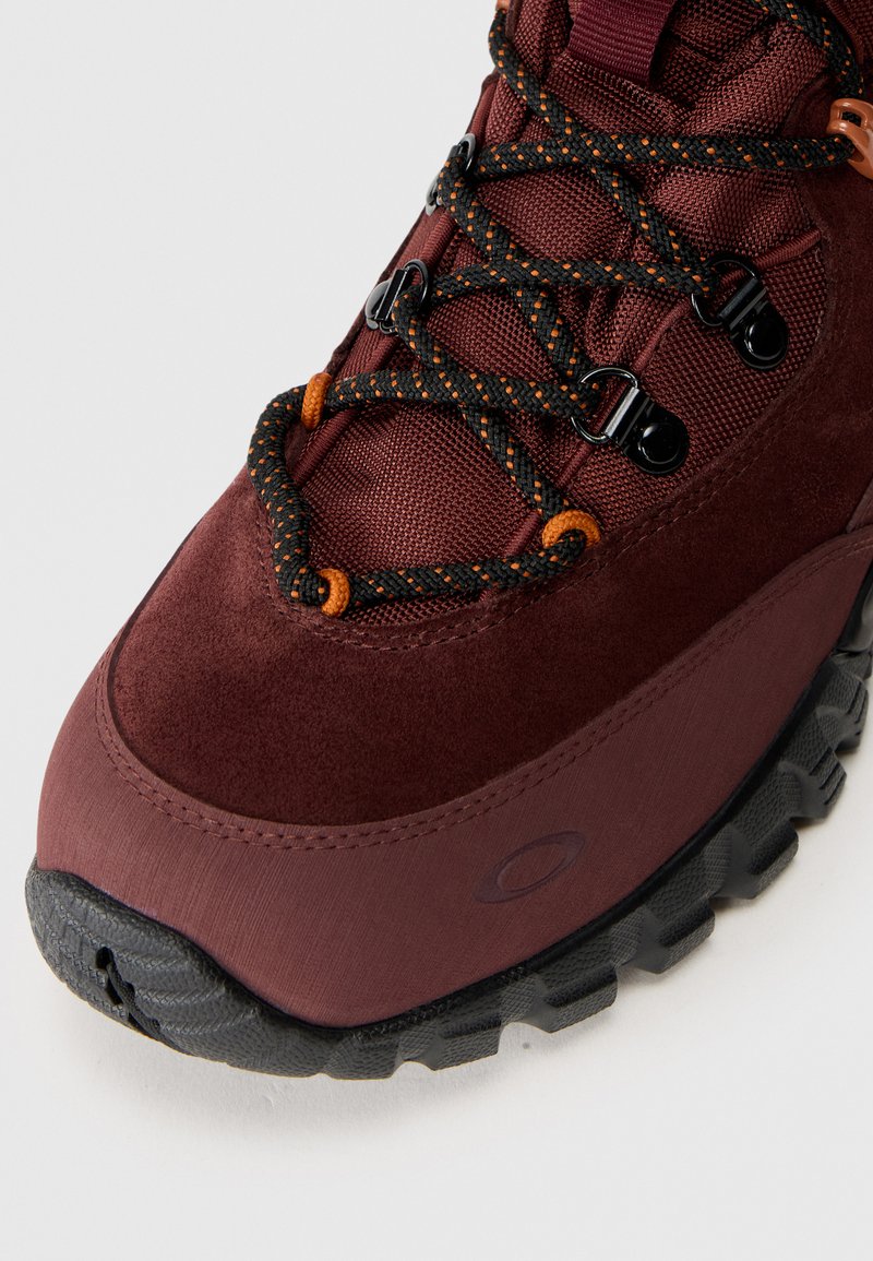 Botas de senderismo en color marrón con parte superior de ante texturizado, cordones negros y naranjas, suela de goma duradera y ojales de metal para soporte.