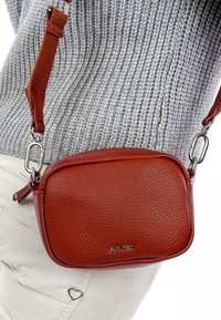 Väike punane tekstureeritud nahast risti kehakott hõbedaste klambrite ja pandlaga, kantud halli kootud kampsuni ja valgete pükstega, millel on südamekujuline ripats.