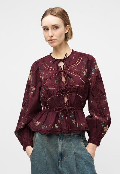Γυναίκα που φοράει μπλούζα μακρυμάνικη βουργουνδί με κεντήματα λουλουδιών, λεπτομέρειες δαντέλας και κλείσιμο με κορδόνια, σε συνδυασμό με ψηλόμεσα μπλε τζιν.