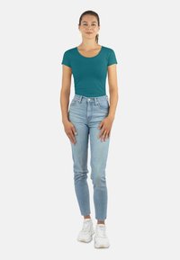 Teal Kurzarmoberteil, kombiniert mit hoch taillierten hellblauen Jeans und weißen Turnschuhen. Das Model steht mit den Händen an den Seiten vor einem einfarbigen Hintergrund.