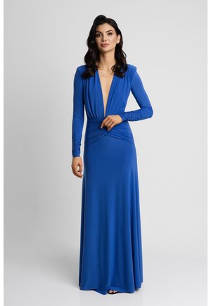 Kobieta o ciemnych włosach, ubrana w długą niebieską suknię z głębokim dekoltem w serek i marszczonymi długimi rękawami, stojąca na jasnym, jednolitym tle.