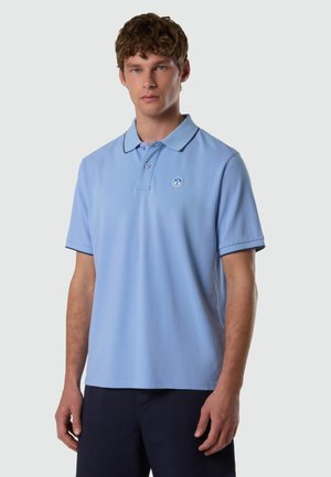 Jeune homme portant un polo bleu clair à manches courtes avec bordure foncée et un pantalon bleu marine, se tenant devant un fond gris uni.