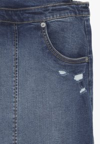 Jeans in denim blu scuro con un aspetto rovinato, caratterizzati da una tasca frontale, cuciture sottili arancioni e orli sfilacciati vicino al ginocchio.