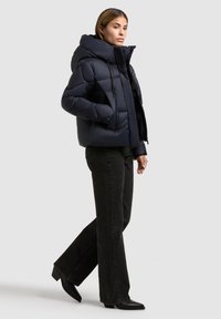Marineblauwe gewatteerde jas met een ruime capuchon, doorgestikte ontwerp en rits aan de voorkant; gecombineerd met zwarte wijd-uitlopende jeans en puntige enkellaarsjes.