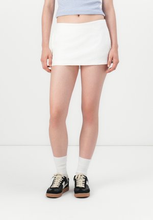 Mini jupe blanche en tissu texturé, associée à des baskets noires et des chaussettes blanches, avec un design minimaliste et une coupe droite.