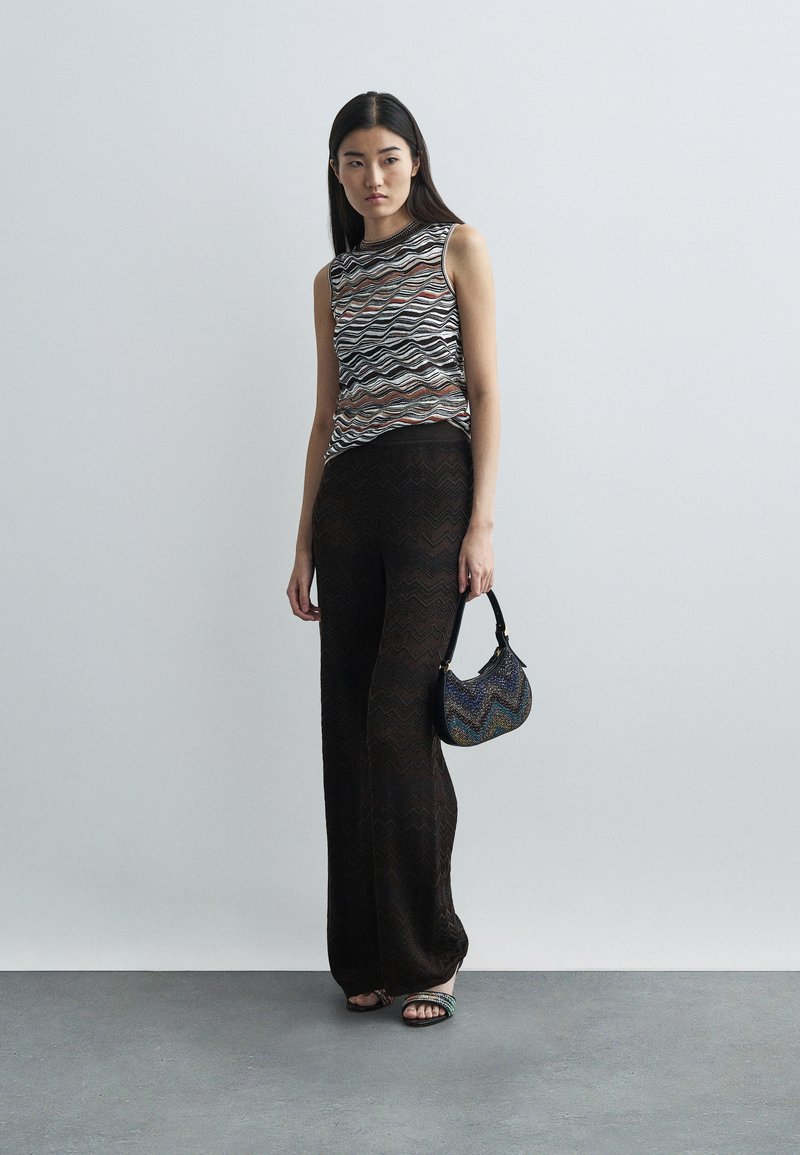 Kvinna i mönstrad ärmlös topp och svarta vida byxor håller en liten färgglad handväska, stående mot en plain grå vägg.