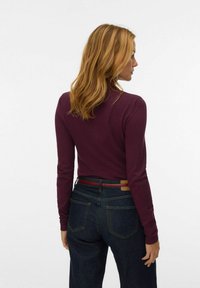 Maglione fitted a maniche lunghe color borgogna con scollatura rotonda, abbinato a jeans in denim scuro con una cintura multicolore e dettagli sulle tasche posteriori.