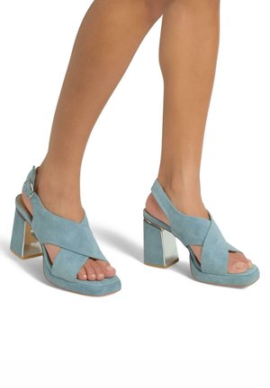 Sandali con tacco in suede color aqua, design con cinghie incrociate, punta aperta e tacco a blocco geometrico specchiato. Texture liscia, forma versatile.