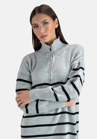 Pull en tricot gris avec des rayures horizontales noires, col montant et fermeture à demi-zip. Dispose de manches longues et de poignets côtelés.