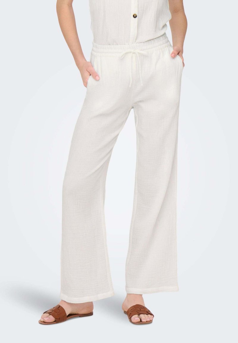 White, lightweight, relaxed-fit pants with a drawstring waist, side pockets, and textured fabric. Paired with brown flat sandals.