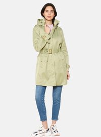 Trinchera impermeable de color verde claro con cintura ceñida, capucha y dos bolsillos frontales. Combinada con jeans ajustados azules y zapatillas blancas.