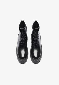 Bottines noires en cuir avec une finition brillante, lacées à l'avant, zips sur les côtés et semelle épaisse. Boucles plates à l'arrière pour un enfilage facile.
