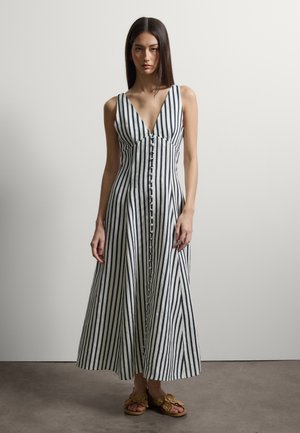 Frau trägt ein ärmelloses Maxikleid mit schwarzen und weißen vertikalen Streifen, tiefem V-Ausschnitt und floralen Sandalen.