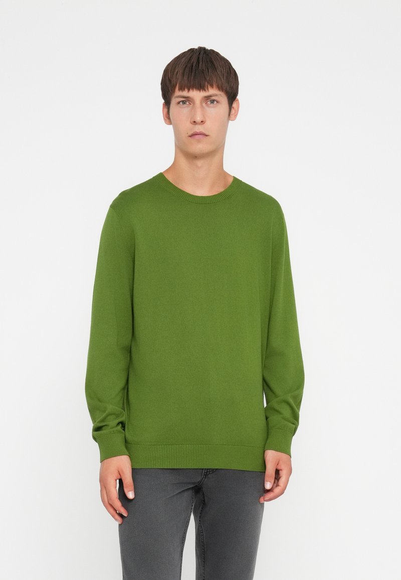 Zöld crewneck pulóver kötött anyagból. Hosszú ujjak ribbelt mandzsettával és hemmeddel. Szürke farmernadrággal viselve. Egyszerű tervezés, nincs minta.