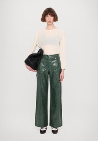 Zwarte geweven tas, groene leren broek met wijde pijpen en een glanzende afwerking, en een doorschijnend crèmekleurig langoverhemd met verticale strepen.