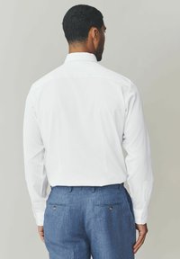 Camicia bianca a maniche lunghe con colletto classico, abbinata a pantaloni blu con trama, tasche posteriori e dettagli con bottoni.