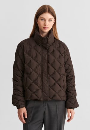 Jeune femme portant une doudoune matelassée brun foncé avec un col haut et un pantalon gris, debout devant un fond clair uni.