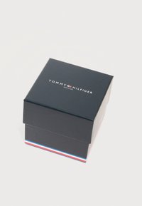 Κλειστό σκούρο μπλε κουτί ρολογιού Tommy Hilfiger με το λογότυπο στο καπάκι και κόκκινες, λευκές και μπλε ρίγες κατά μήκος της κάτω ακμής, σε λευκό φόντο.