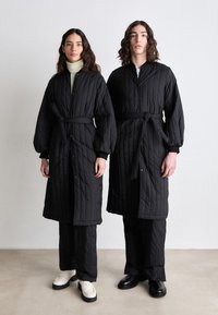Black quilted coats with a belted waist and snap closures, paired with black pants and contrasting shoes—one white, one black.