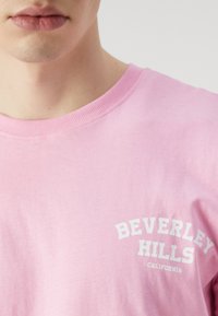 Rožinė medvilninė marškinė su apvaliu iškirpimu ir trumpomis rankovėmis. Ant jų yra balti žodžiai „BEVERLEY HILLS KALIFORNIJA“ išlenktame dizaine.