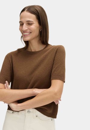 Uśmiechnięta kobieta o długich brązowych włosach, ubrana w brązową, krótkorękawową dzianinową bluzkę i jasne spodnie, z założonymi rękami, zamkniętymi oczami, zwrócona lekko w lewo.