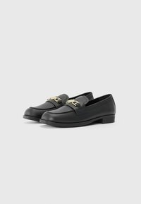 Sapatos mocassim de couro preto com textura suave, salto bloco baixo e um detalhe em corrente dourada na cinta da frente. Design simples e moderno.