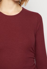 Burgundy långärmad topp med ribbat mönster, rund halsringning och samlat detaljer på sidan, som visar en åtsittande silhuett.