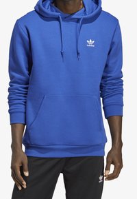 Sininen vetoketjullinen huppari pehmeästä kankaasta, jossa on etutasku, ribbireunat, säädettävä nyöri ja valkoinen logo rinnassa.