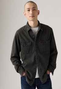 Tmavosivá košeľa na gombíky s dvoma náprsnými vreckami, golierom a dlhými rukávmi, doplnená bielym tričkom a tmavomodrými džínsami.