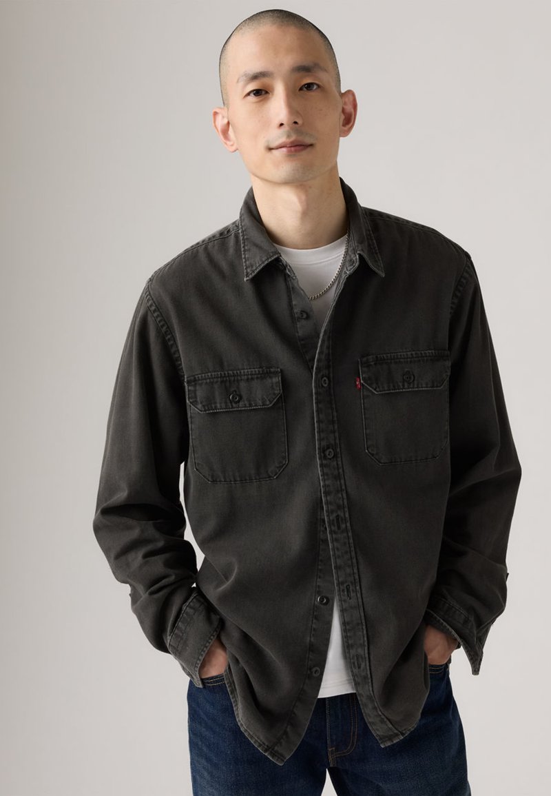 Chemise boutonnée gris foncé avec deux poches poitrine, un col et des manches longues, associée à un t-shirt blanc et un jean bleu foncé.