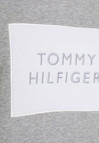 Gray cotton sweatshirt with a white rectangular patch featuring "TOMMY HILFIGER" embroidered in silver. Soft texture, casual design.