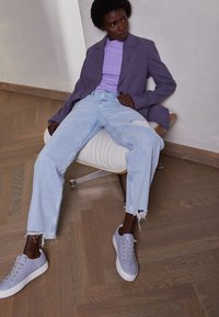 Donna con pelle scura e capelli corti seduta su una sedia bianca, indossando un blazer a quadri viola, una maglietta lavanda, jeans chiari strappati e sneakers di un leggero colore viola.