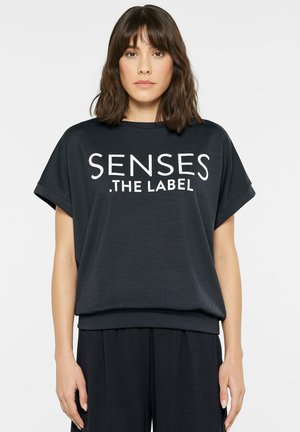 Woman with shoulder-length brown hair wears a black shirt with rolled sleeves displaying the text "SENSES THE LABEL" in white letters.