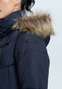 Marine blauwe pufferjack met een afneembare capuchon van pels, featuring een zwart logo-embleem op de mouw en een textiel voor isolatie.