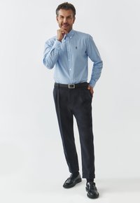 Camisa de cuadros azul y blanca, de botones, de mangas largas, combinada con pantalones oscuros y mocasines negros, en pose de pie con la mano en la barbilla.
