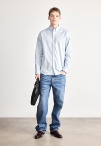 Gaiši zila pogājama krekla, ikdienas zilas džinsa bikses, tumši brūnas kurpes, tur melnu ādas somu, stāv pret neitrālu fonu.