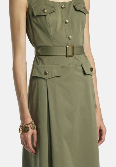 Olīvzaļš kleita bez piedurknēm ar aizpogājamām kabatām ar atloku, atbilstošu jostu ar misiņa sprādzi un zelta monētas rotas aproci uz kreisās rokas plaukstas.