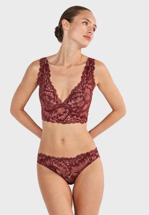 Femme portant un bralette en dentelle bordeaux assorti et un shorty, debout, les mains dans le dos, regardant sur le côté sur un fond uni.