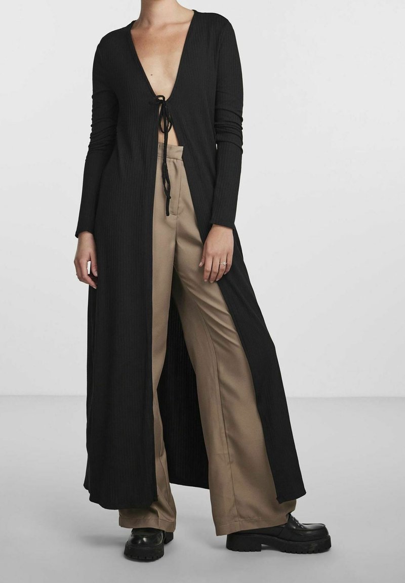 Femme portant un long cardigan noir ouvert devant, noué à la poitrine, pantalon beige à jambes larges et chaussures compensées noires sur fond blanc.